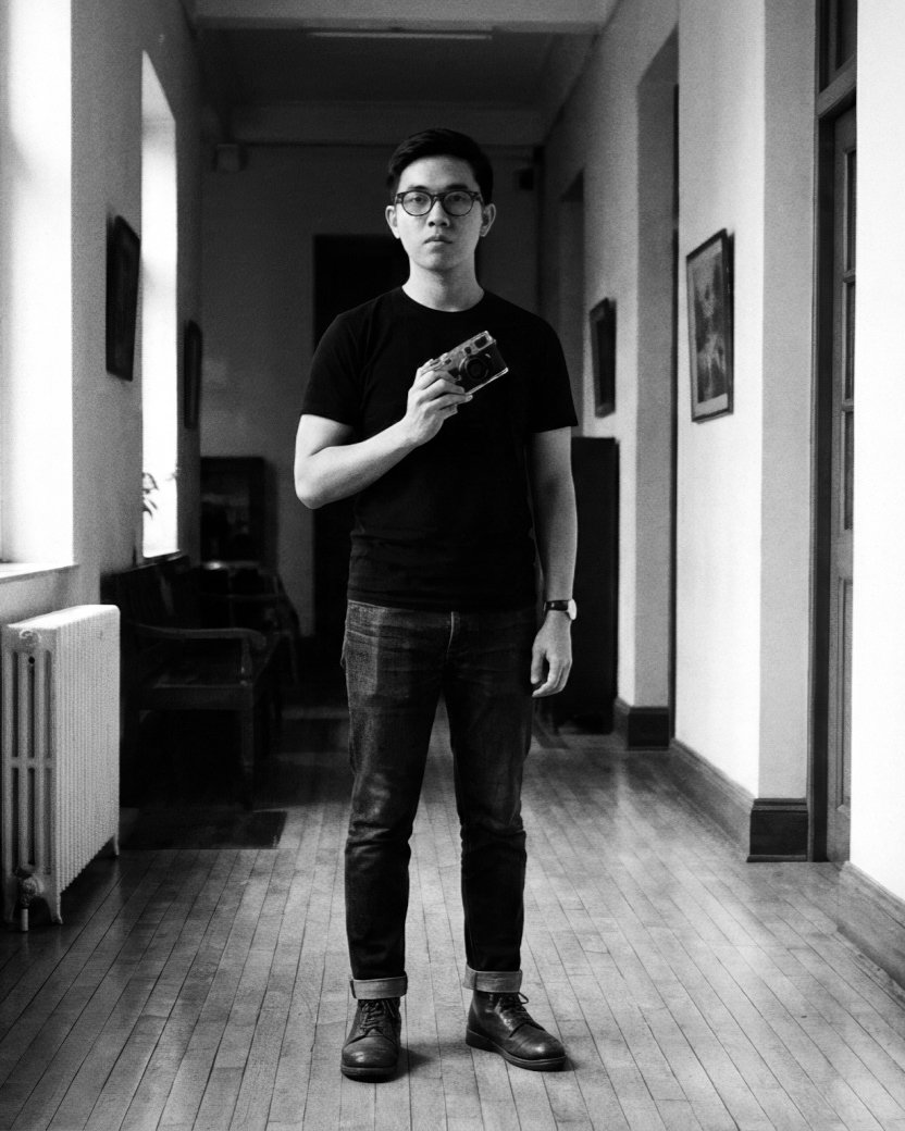 Portrait of Minh Trần in a hallway, holding a Leica rangefinder — the tool of the trade.
